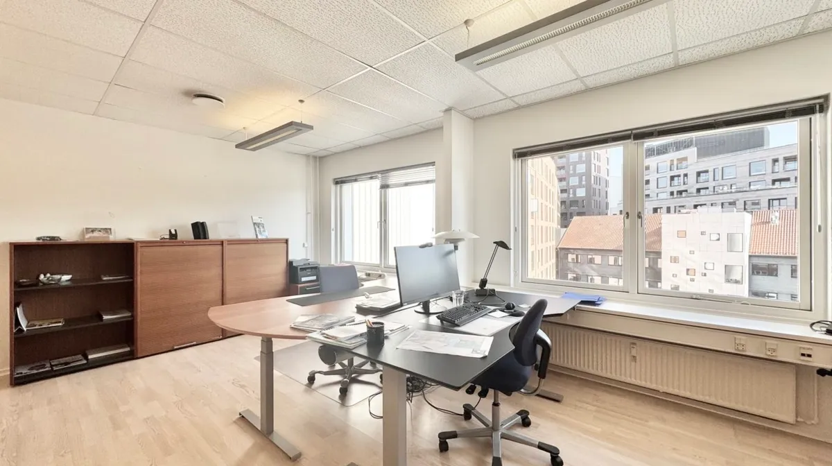 Et kontorlokale med vinduer, hvor man kan se bygninger udenfor. Et bord med en computer og en stol står mod vinduet. Et skab