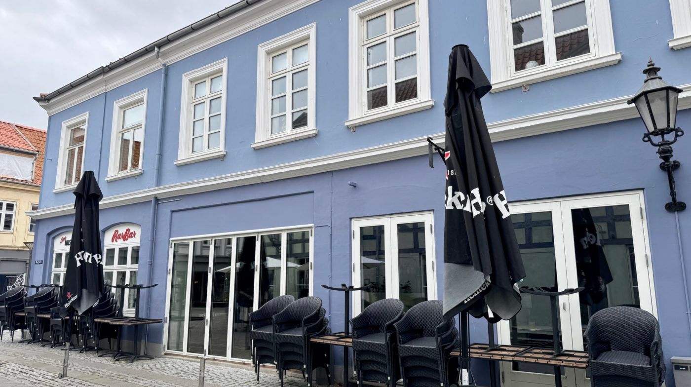 Restaurant med blå væg og hvide ruder, glasdøre, terrassestole og parasoller i sort. Lampestøtter og bord på gaden foran