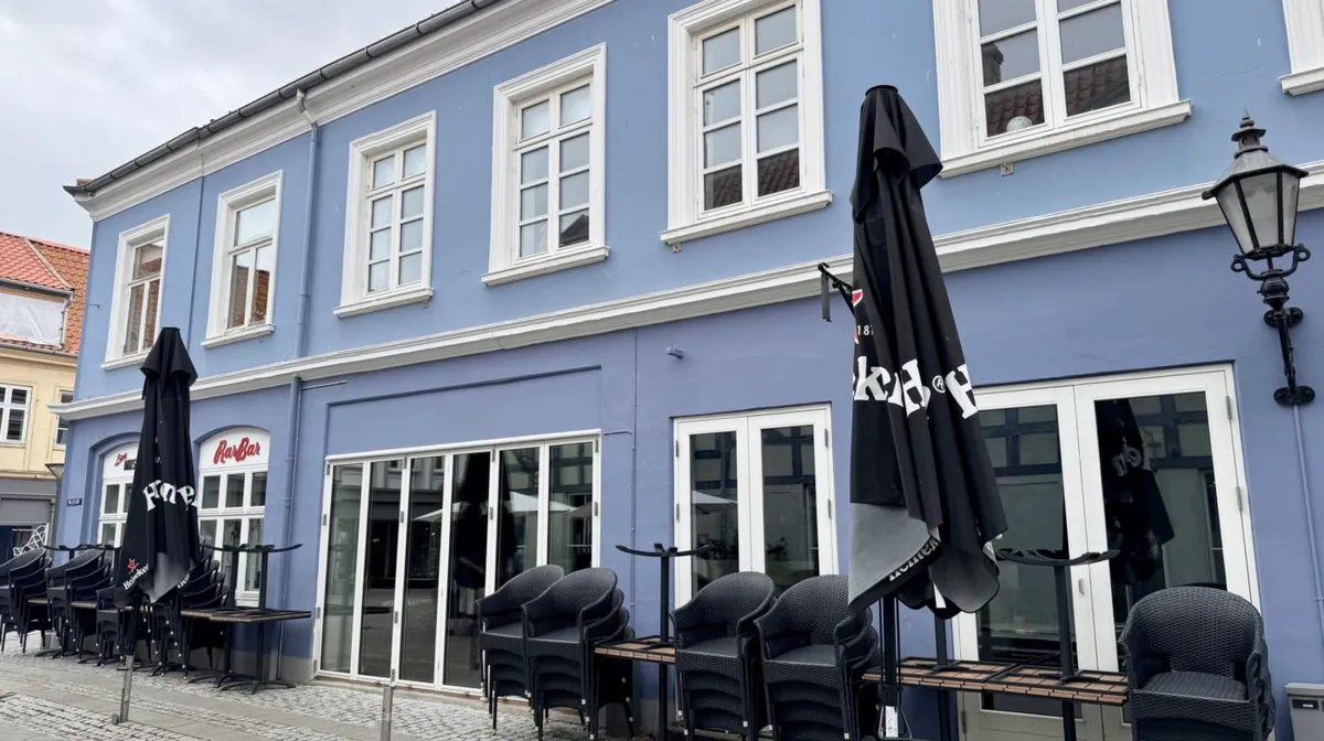 Restaurant med blå væg og hvide ruder, glasdøre, terrassestole og parasoller i sort. Lampestøtter og bord på gaden foran