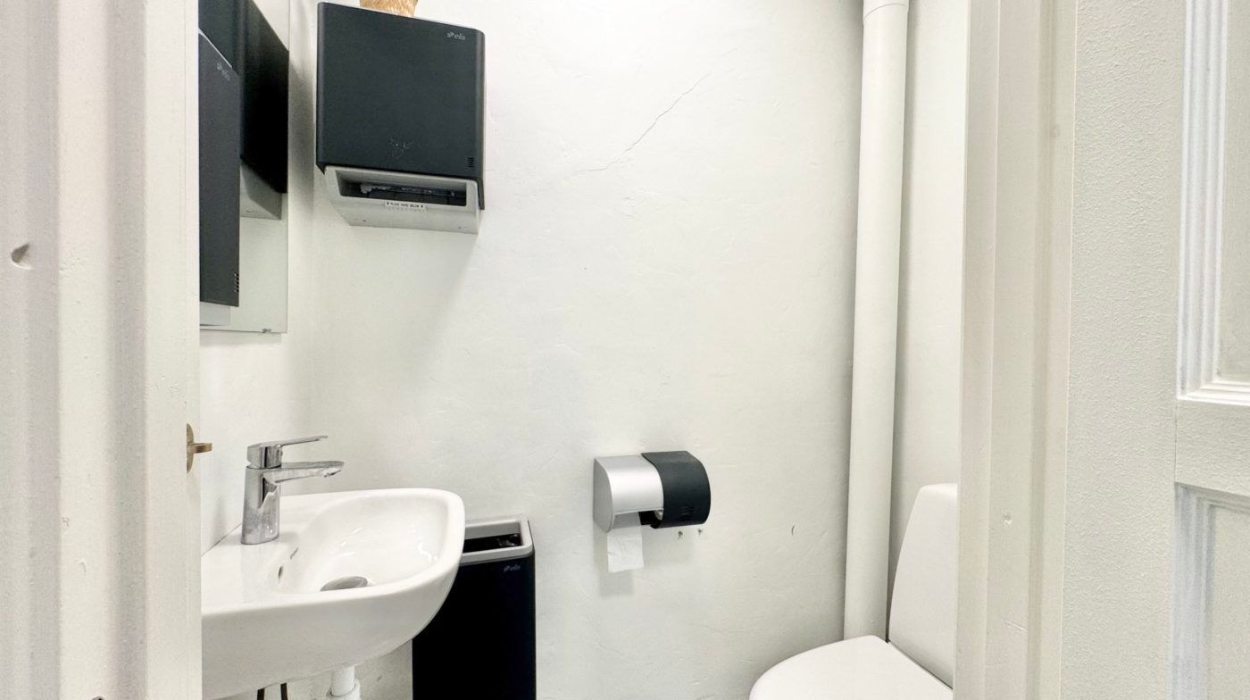 Foto af et lille badeværelse med hvidt toilet og hvidt vask, toiletpapirhylde med en rull papir og en hvid spejl med en