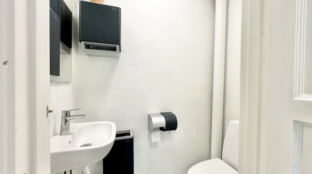 Foto af et lille badeværelse med hvidt toilet og hvidt vask, toiletpapirhylde med en rull papir og en hvid spejl med en