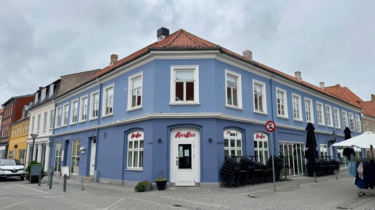 Nyborg Centrum byder på en hjørnebygning med blå facade og røde tagplader. En bilparkeret foran og to paraplyer med klæder i