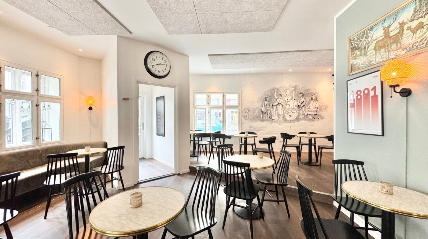 En indre café med høje, brede vinduer, gulvtæppe, møbler og kunstværker på væggen, samt lyse gulvlampe og et ur på væggen