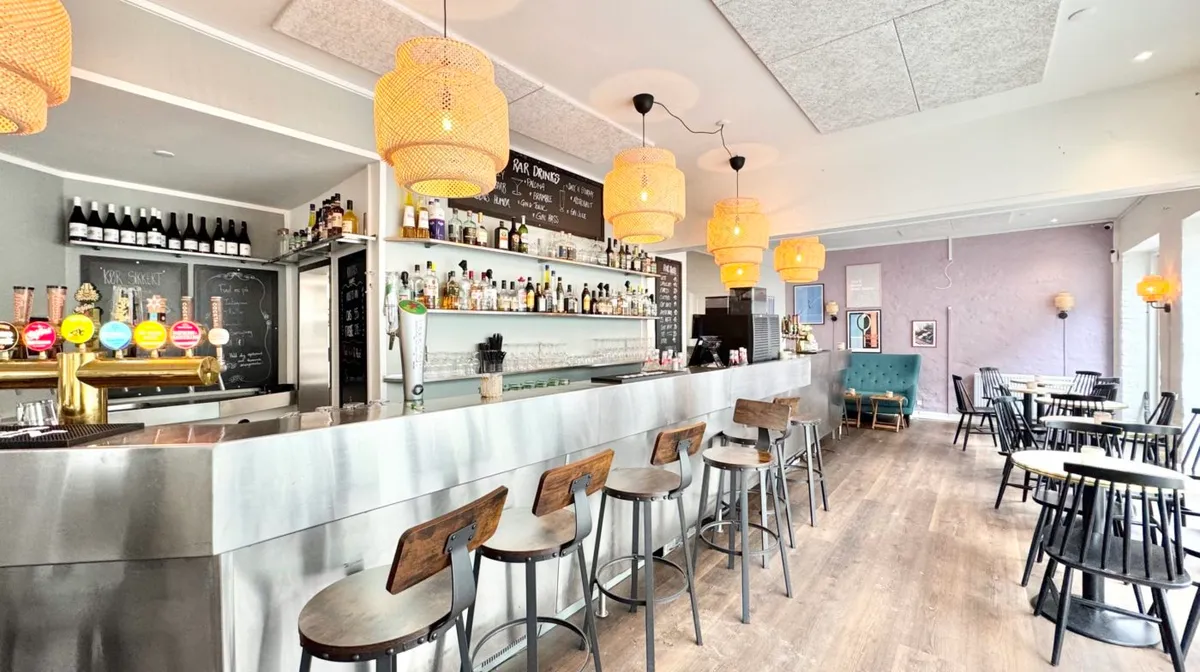 Inde i en hyggelig restaurant med møbler i træ og metal, bar med champagner og cocktailglas, og en sort tavle med menuen