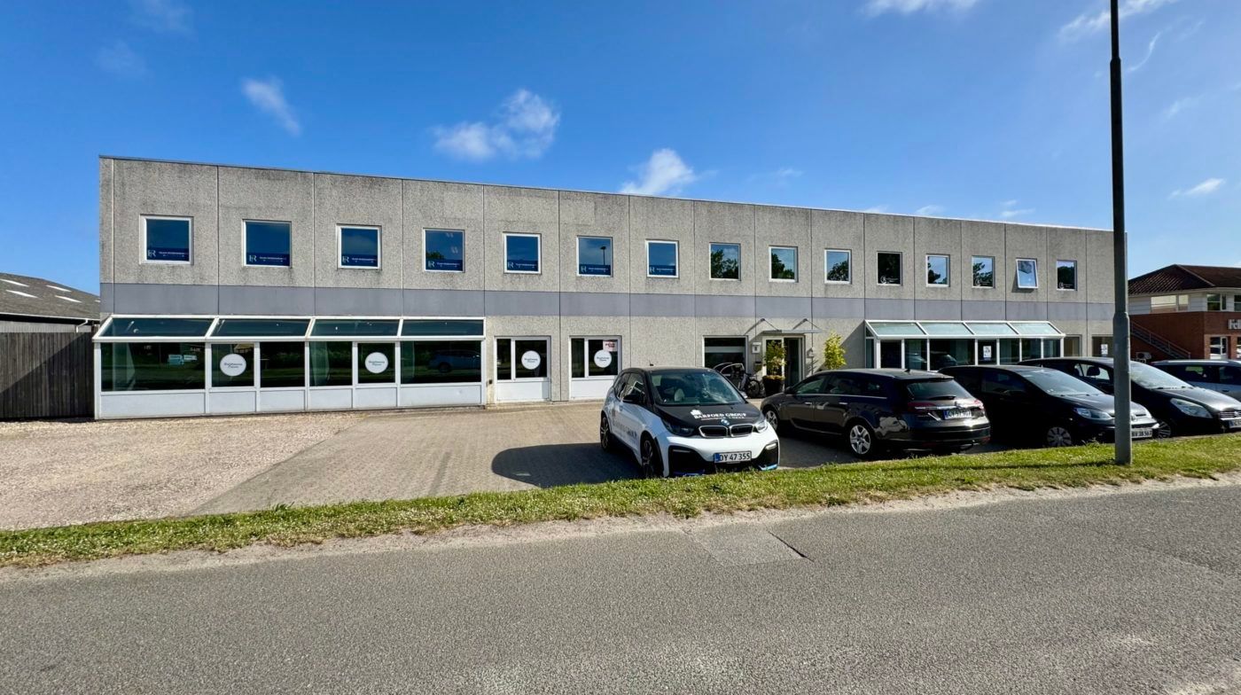 Foto af en stor kontorbygning med store vinduer og flere parkerede biler foran og bagved bygningen