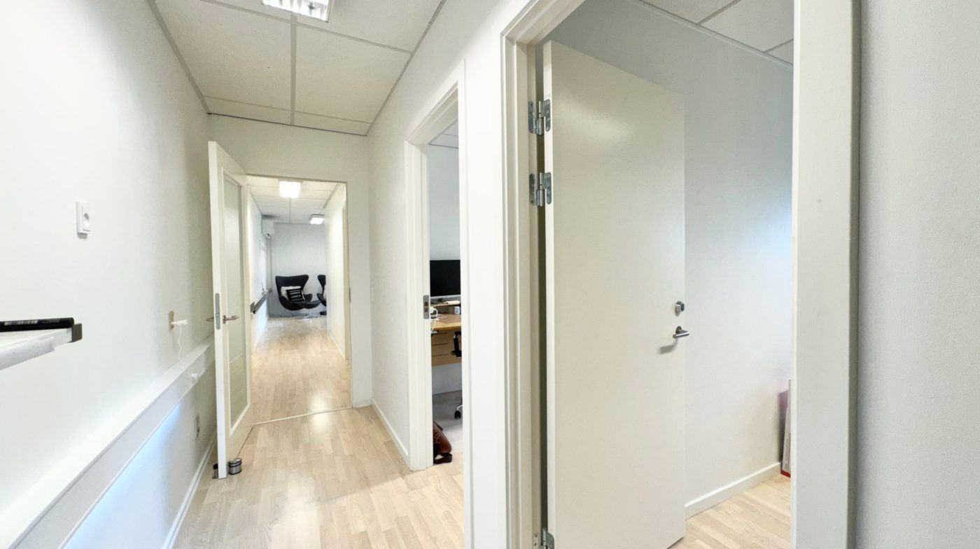 Køkken med åben dør og lyse lofter og højrekants kontordør. Værelse med bord, computer og stol samt toilet.