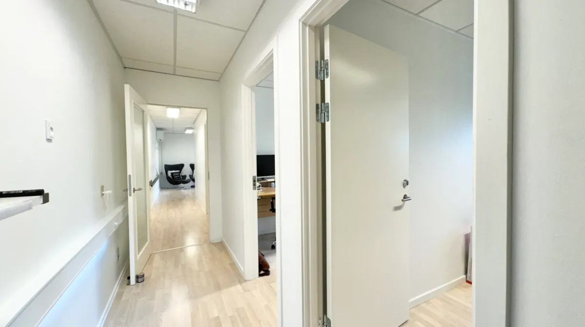 Køkken med åben dør og lyse lofter og højrekants kontordør. Værelse med bord, computer og stol samt toilet.