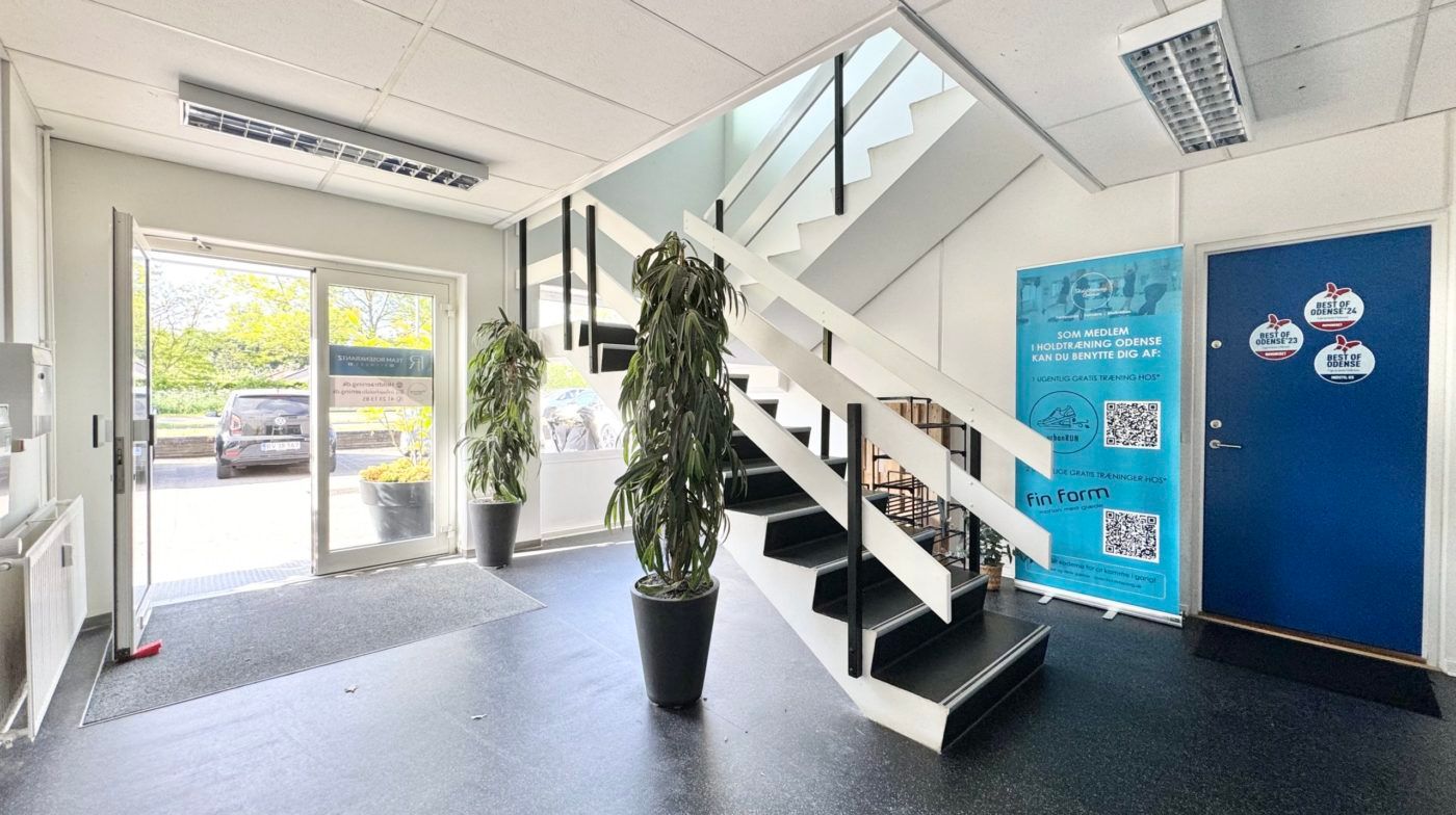 Indvendigt i lokaler er der en åben dør, hvorfra man ser ud på en parkeringsplads med biler. Stående er en plante og en