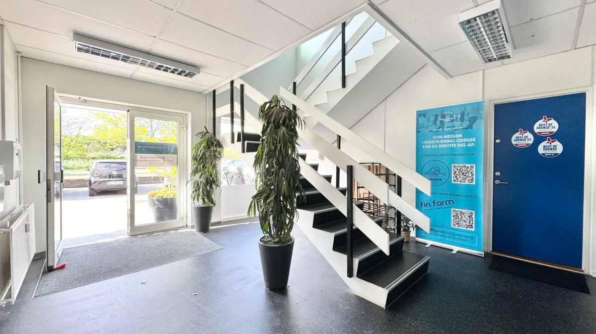 Indvendigt i lokaler er der en åben dør, hvorfra man ser ud på en parkeringsplads med biler. Stående er en plante og en