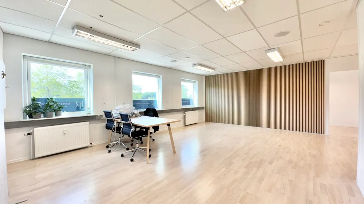 Lyst kontor med højt loft og hvid tapet. Trægulv, lysevinduer, radiator og plantepoter. Et bord med fire stole.