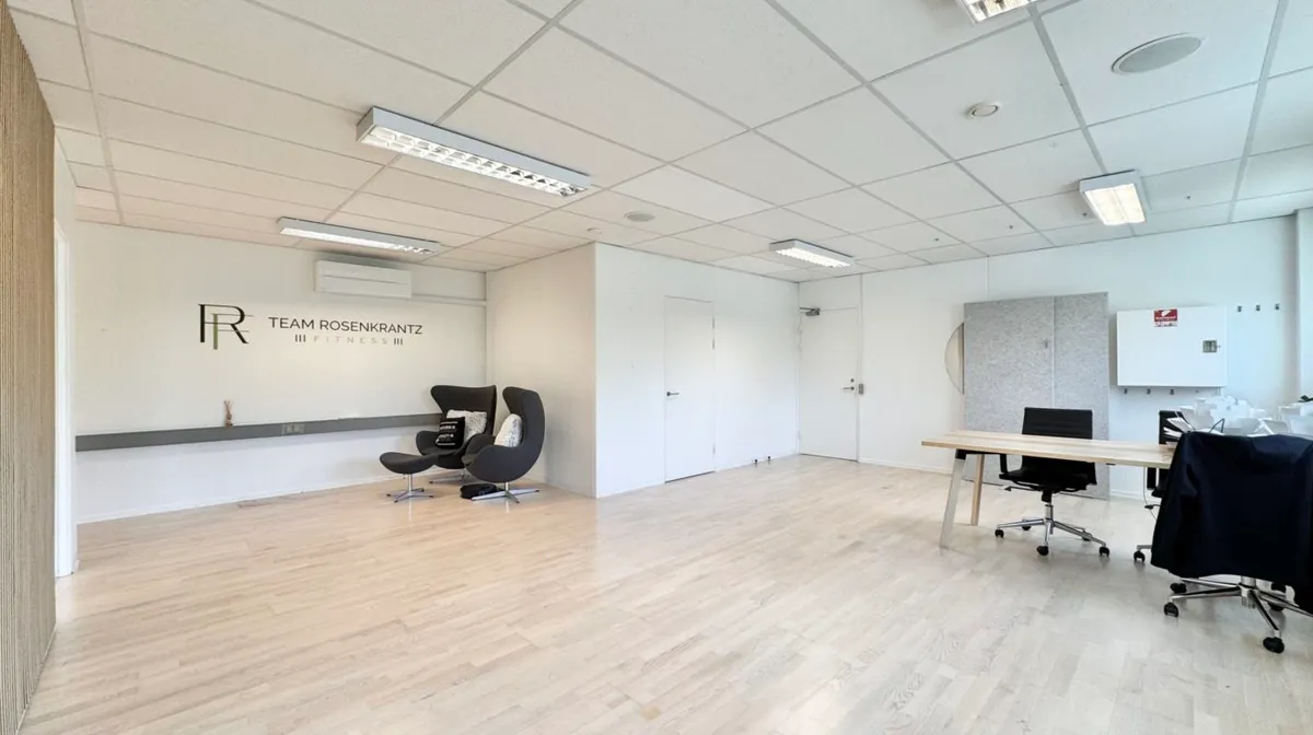 Lyst og funktionelt kontorrum med lyse trægulve og lyse loftslampe. To stole og to skriveborde med høje stole og en sofa i