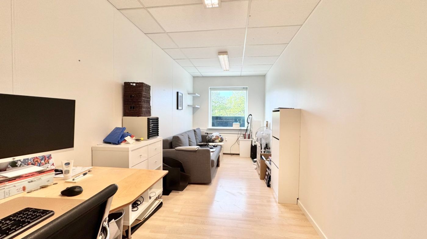 Lys kontor med bord, stol, skrivebord, skærm, sofa og hylder samt vindue med udsigt til grønne træer