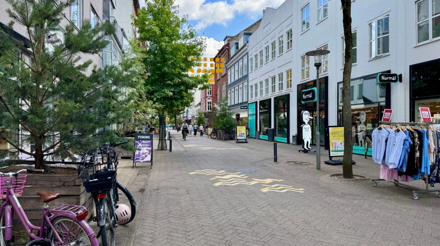 Butiksgade med træer og cykler og reklameplakater samt midtvejssalg og midtsommersalg