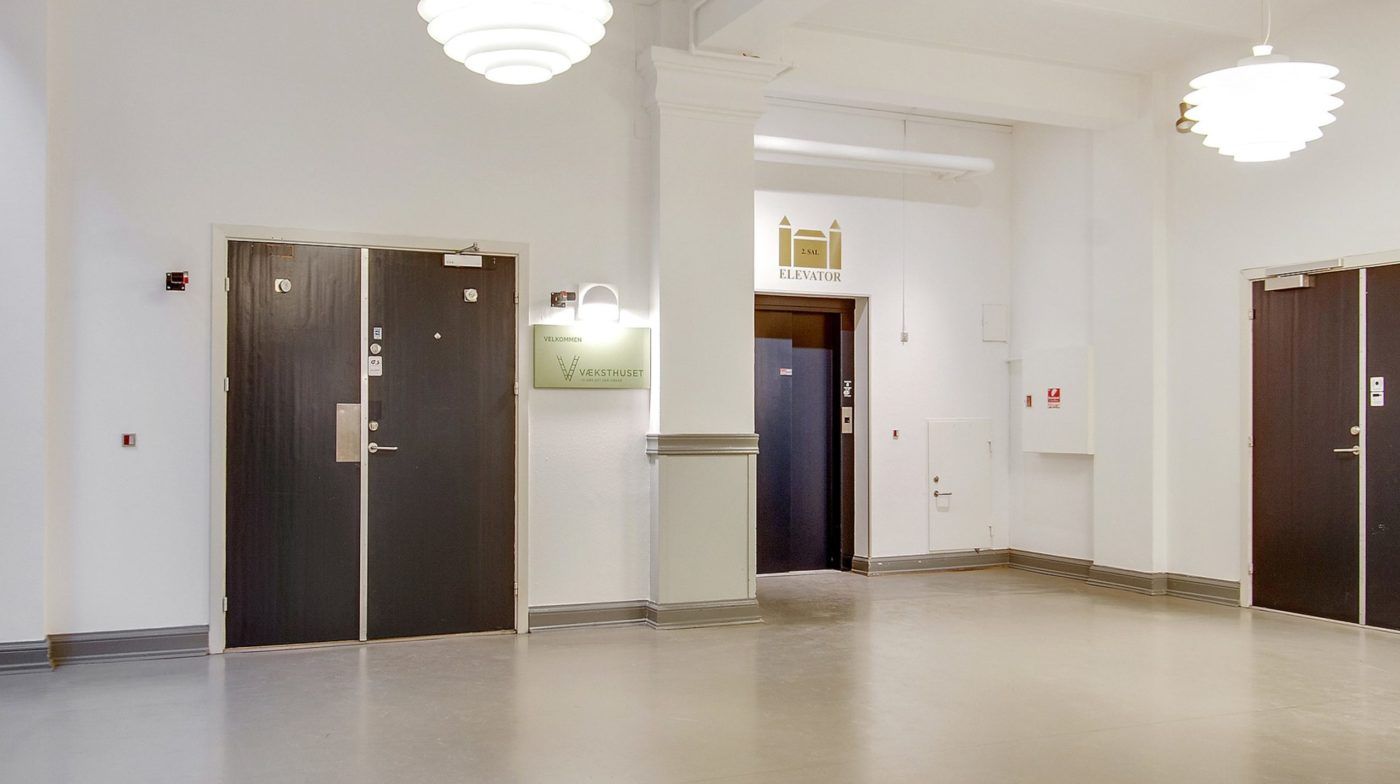 En lys hall med to dobbeltdøre og en elevator. Der er et skilt med teksten "Vælkommen" og et elevatorskilt.