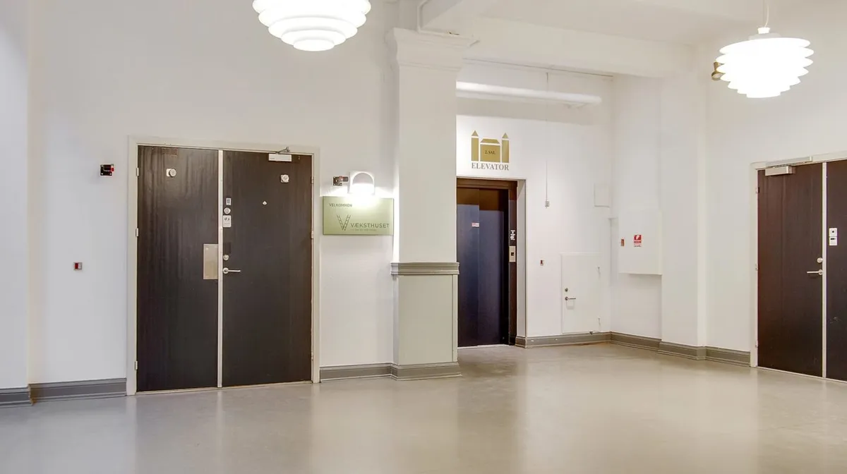 En lys hall med to dobbeltdøre og en elevator. Der er et skilt med teksten "Vælkommen" og et elevatorskilt.