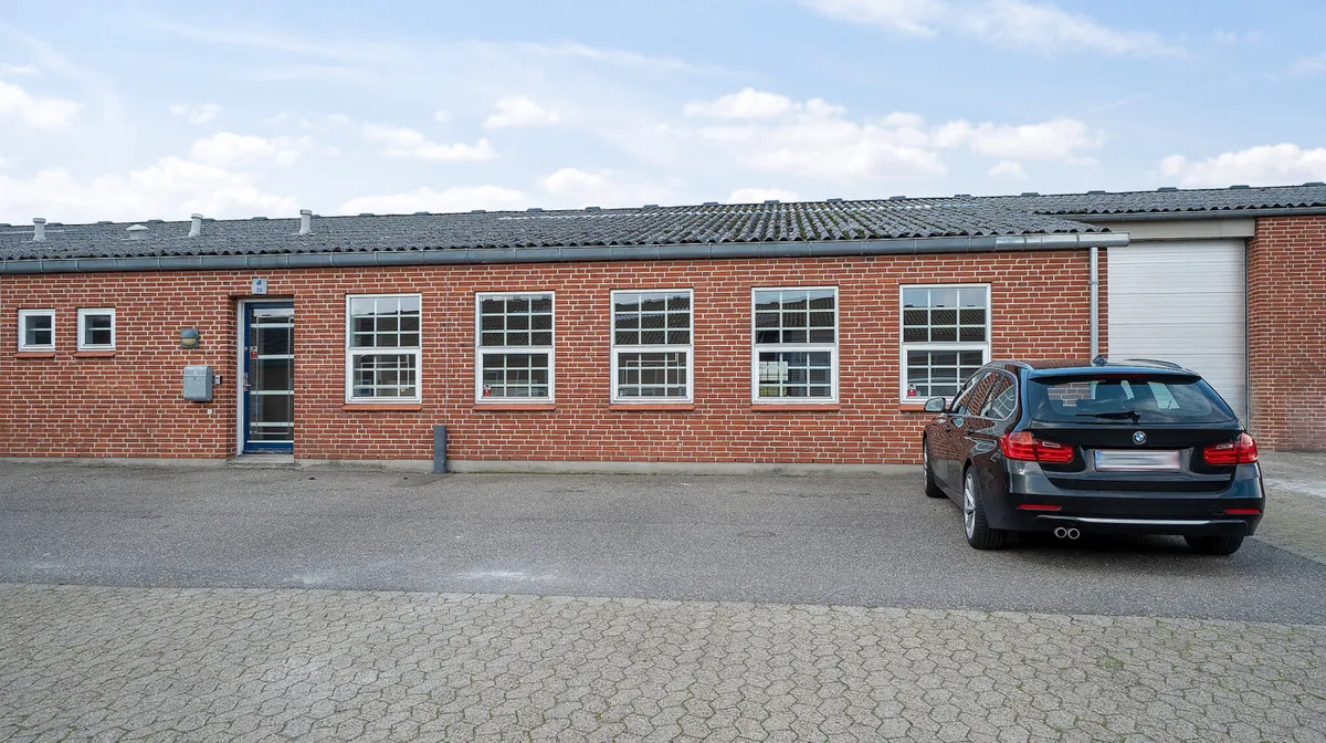 Et rødt murstensbygning med flere vinduer og et garagedør i højre side. En sort BMW parkeret foran bygningen.