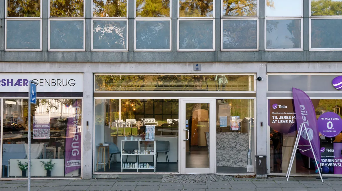 Butiksfront med skilte og reklamebannere, søjle med handicaptegn og Telia-banner samt indvendige butiksvinduer med produkter