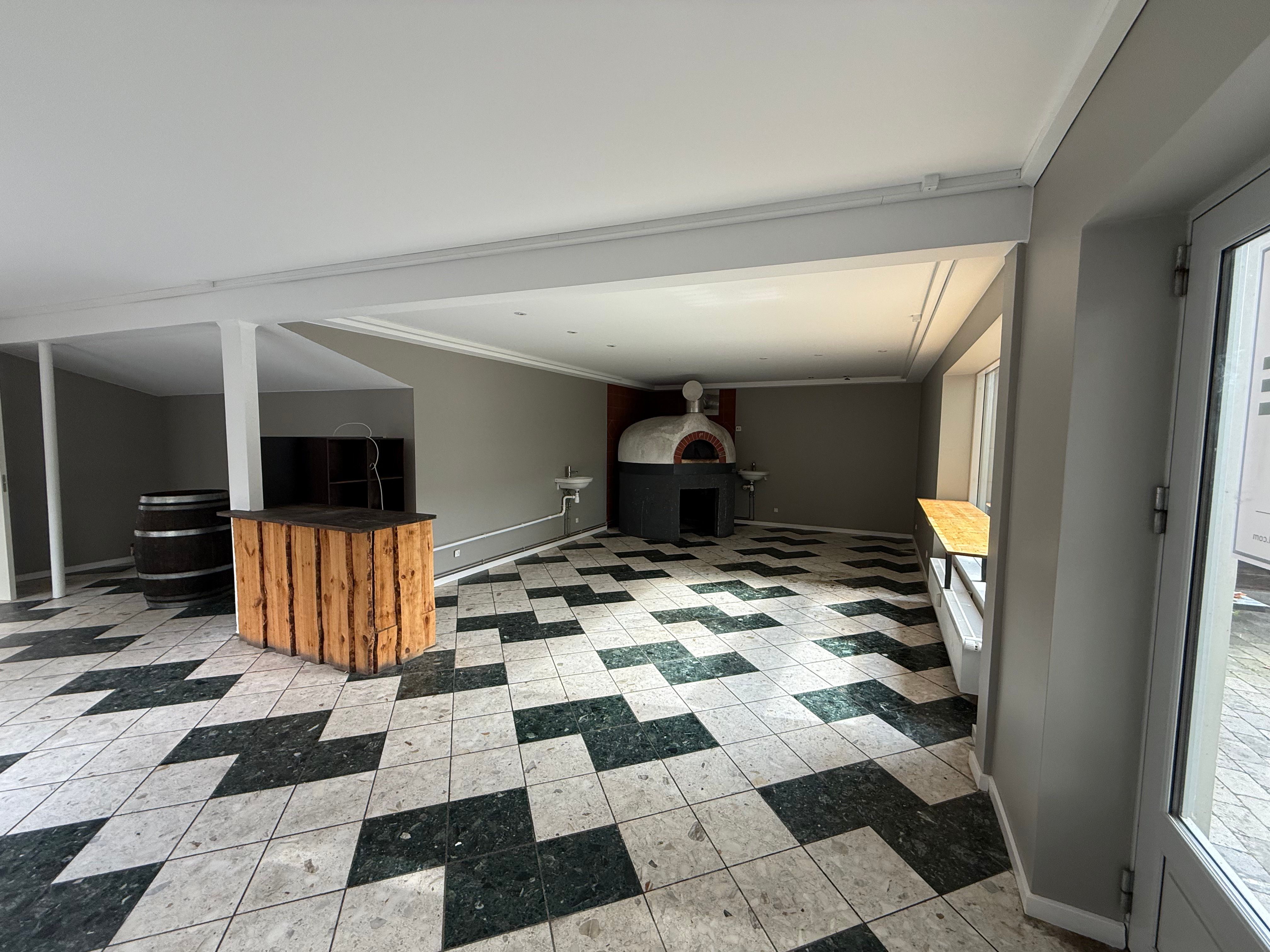 En tom lokale med tønde, træbord, pizzaovn og søjler med to døre og vinkler i væggen