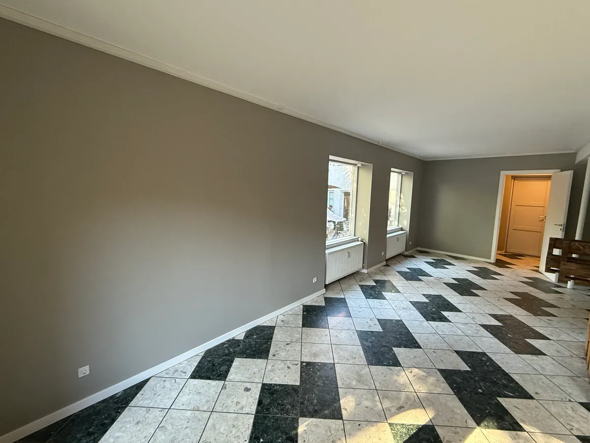 En tom lejlighed med lyse og mørke kvadratiske plader på gulvet og lysegrå vægge samt to vinduer med lysegrå ruder og hvide