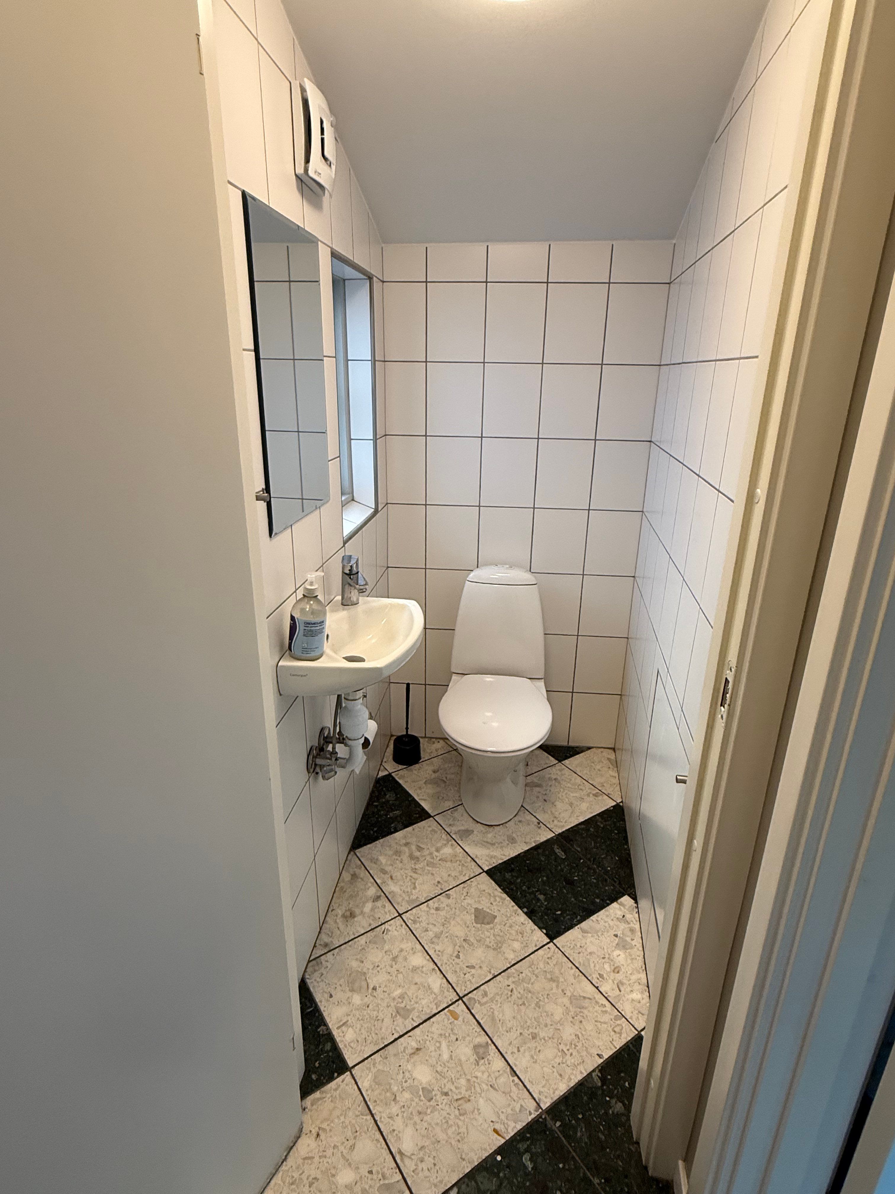 En lille toiletrum med hvide vægge og møbler, hvor der er en hvid toilet, et hvidt toiletbord og en hvid spejl med en flaske