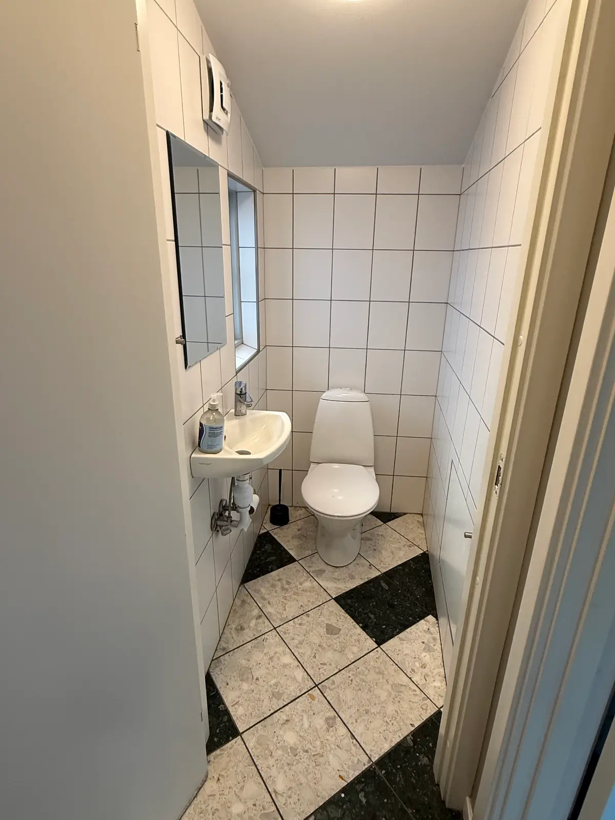 En lille toiletrum med hvide vægge og møbler, hvor der er en hvid toilet, et hvidt toiletbord og en hvid spejl med en flaske
