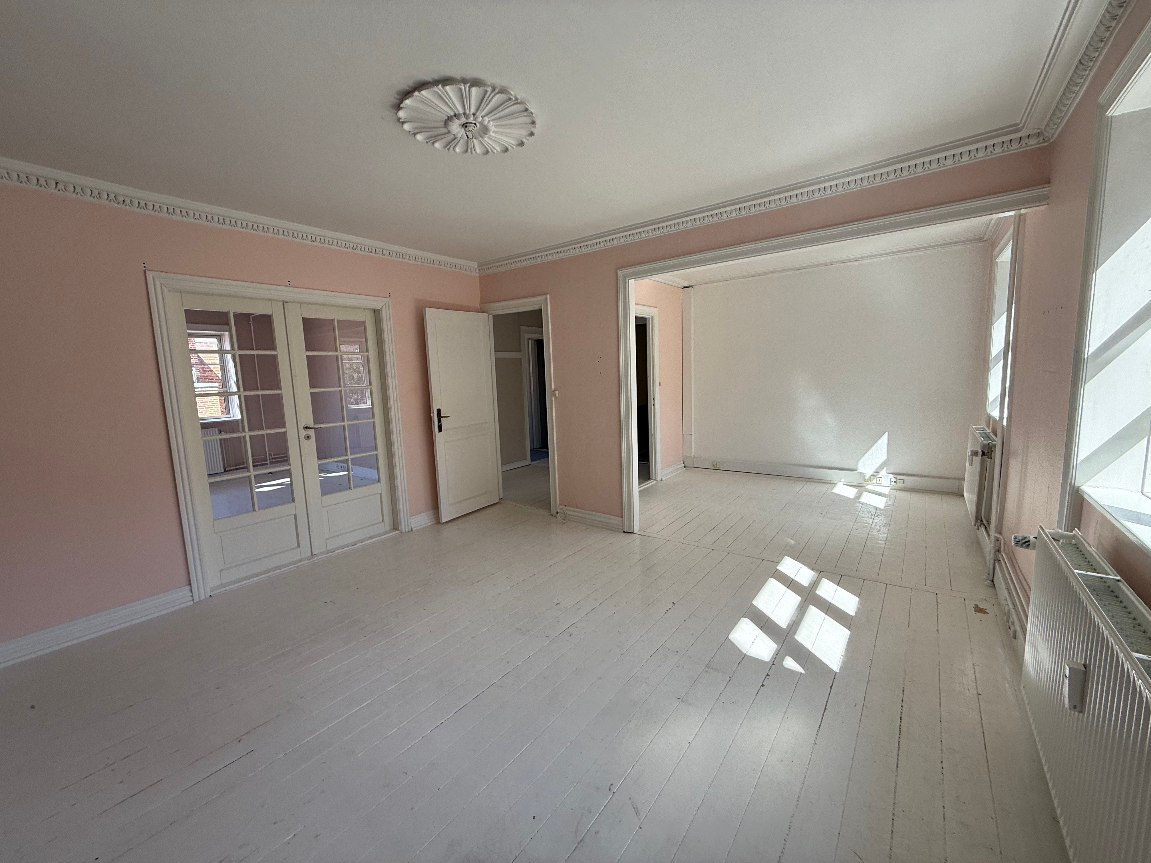 En stor, tom stue med hvide gulvplanker og lyse vægge med vindue og radiatorer