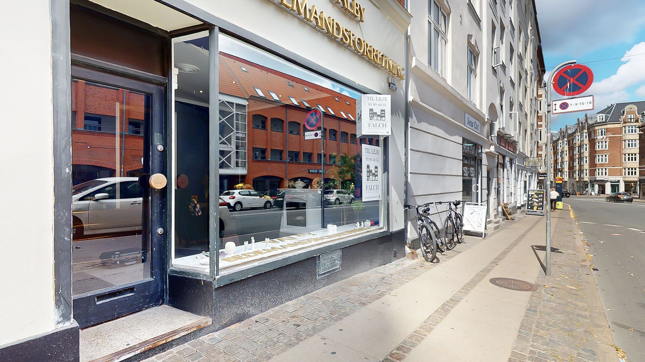 Storefront med vinduer og plakater omkring åbningstider og adresser. To cykler parkeret på siden af gaden og en mand står