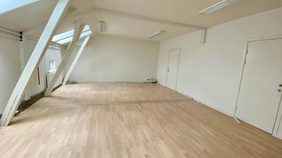 Et stort og lyst værelse med lysegule trægulve, hvide vægge og loftsrum med loftsvinduer og hvide bærende bjælker