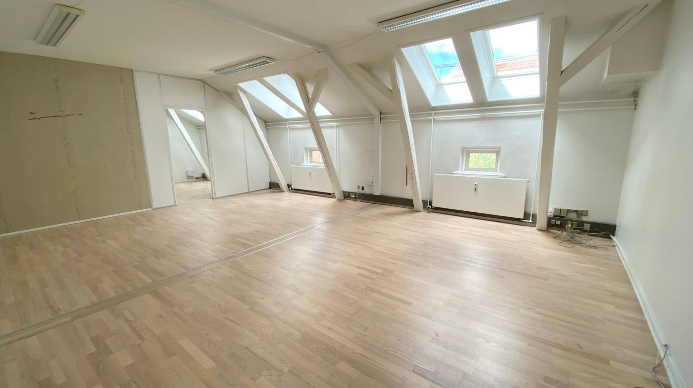 To lyse loftsrum med lysegult trægulv, hvide vægge og loftsvinduer samt en hvid radiator i enden af rummet