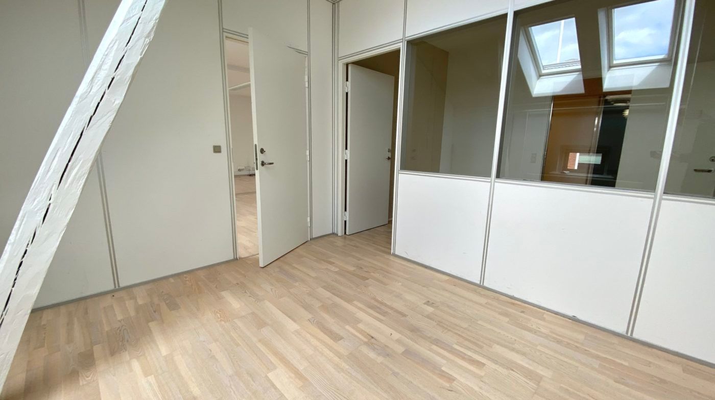 Et lyst og åbent kontorrum med trægulve og hvide vægge og lofter, der afsluttes med en hvid loftsleje med lys.