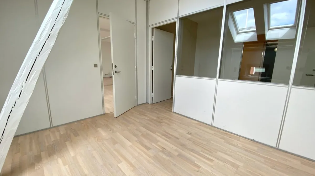 Et lyst og åbent kontorrum med trægulve og hvide vægge og lofter, der afsluttes med en hvid loftsleje med lys.