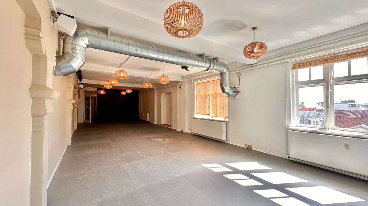 En stor, tom hall med høje hvide vægge, lyse trægulve og store vinduer med skurer samt lysekroner hængende fra loftet