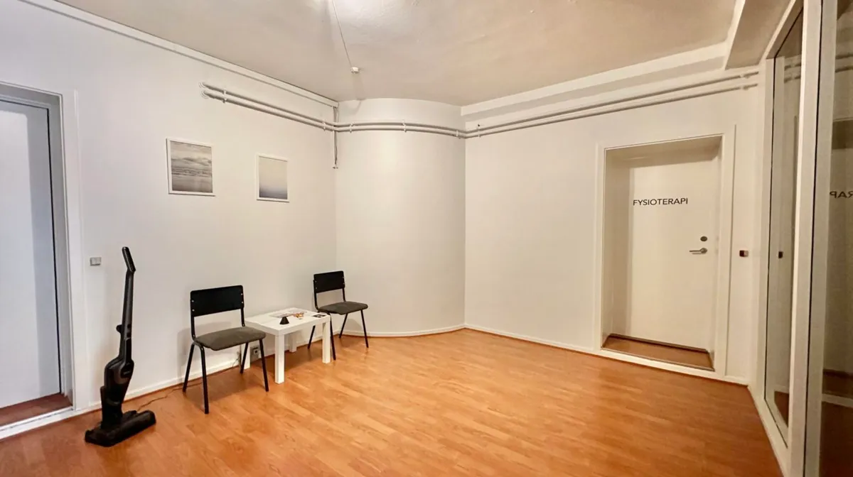 Et stort værelse med to stole og et bord, samt en støvsuger. Værelset har hvide vægge og lyse trægulve