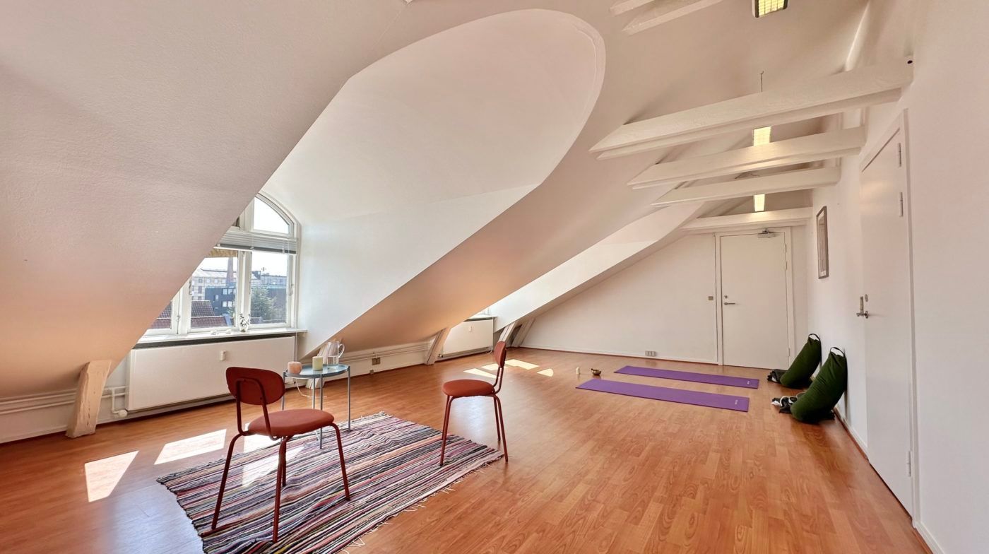 Yogamatte og yogakudde på loftet. To stole med søjler og et lille bord med to stearinlys. Lyse loftsbjælke og to hvide døre