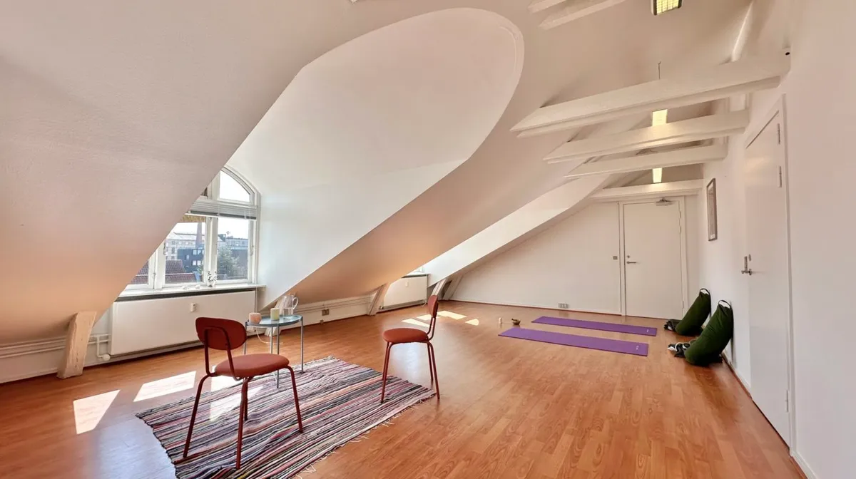 Yogamatte og yogakudde på loftet. To stole med søjler og et lille bord med to stearinlys. Lyse loftsbjælke og to hvide døre