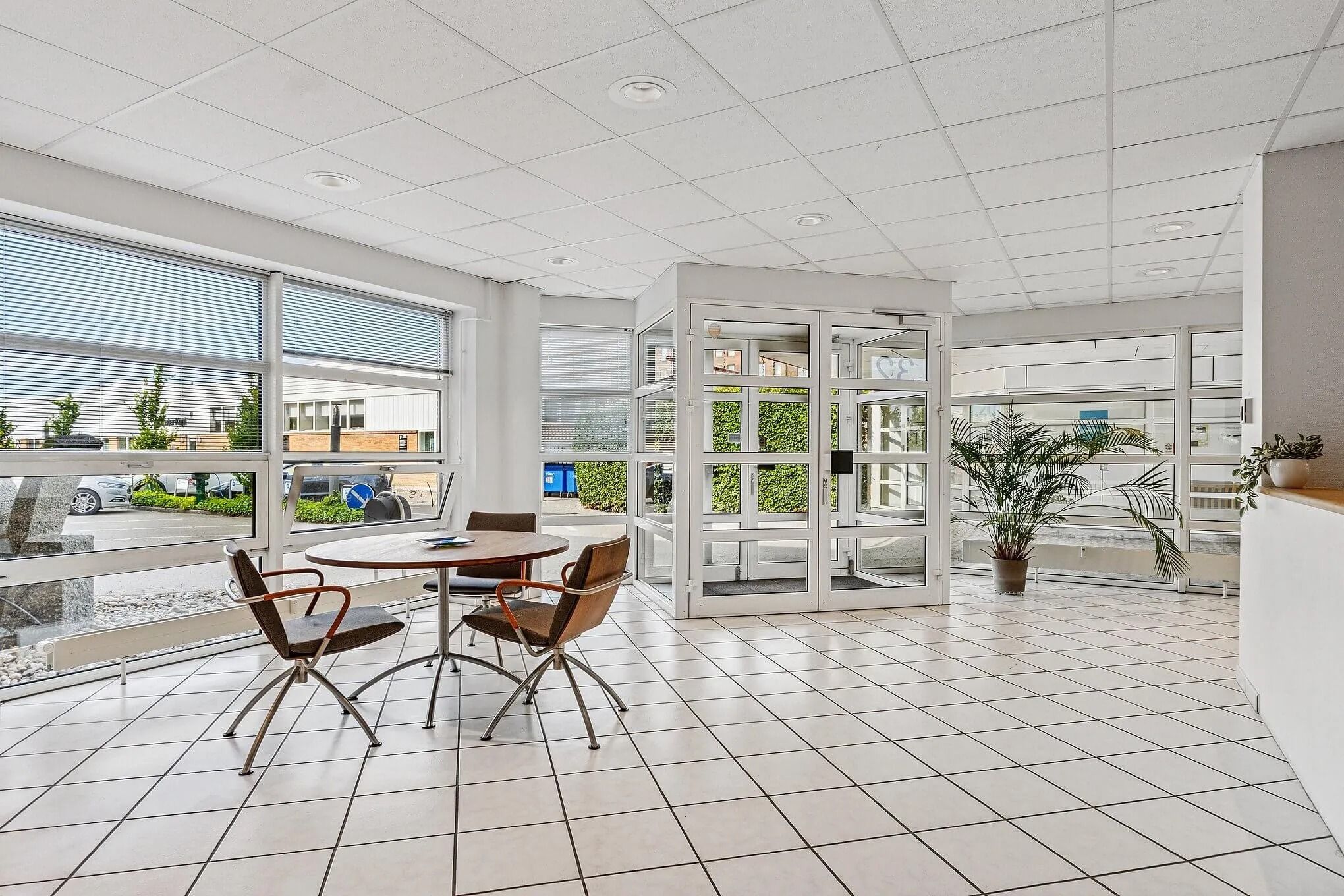 Kontor med moderne møbler og plante, hvide vægge og gulv samt store glasruder med udsigt til parkeringsplads og bygninger