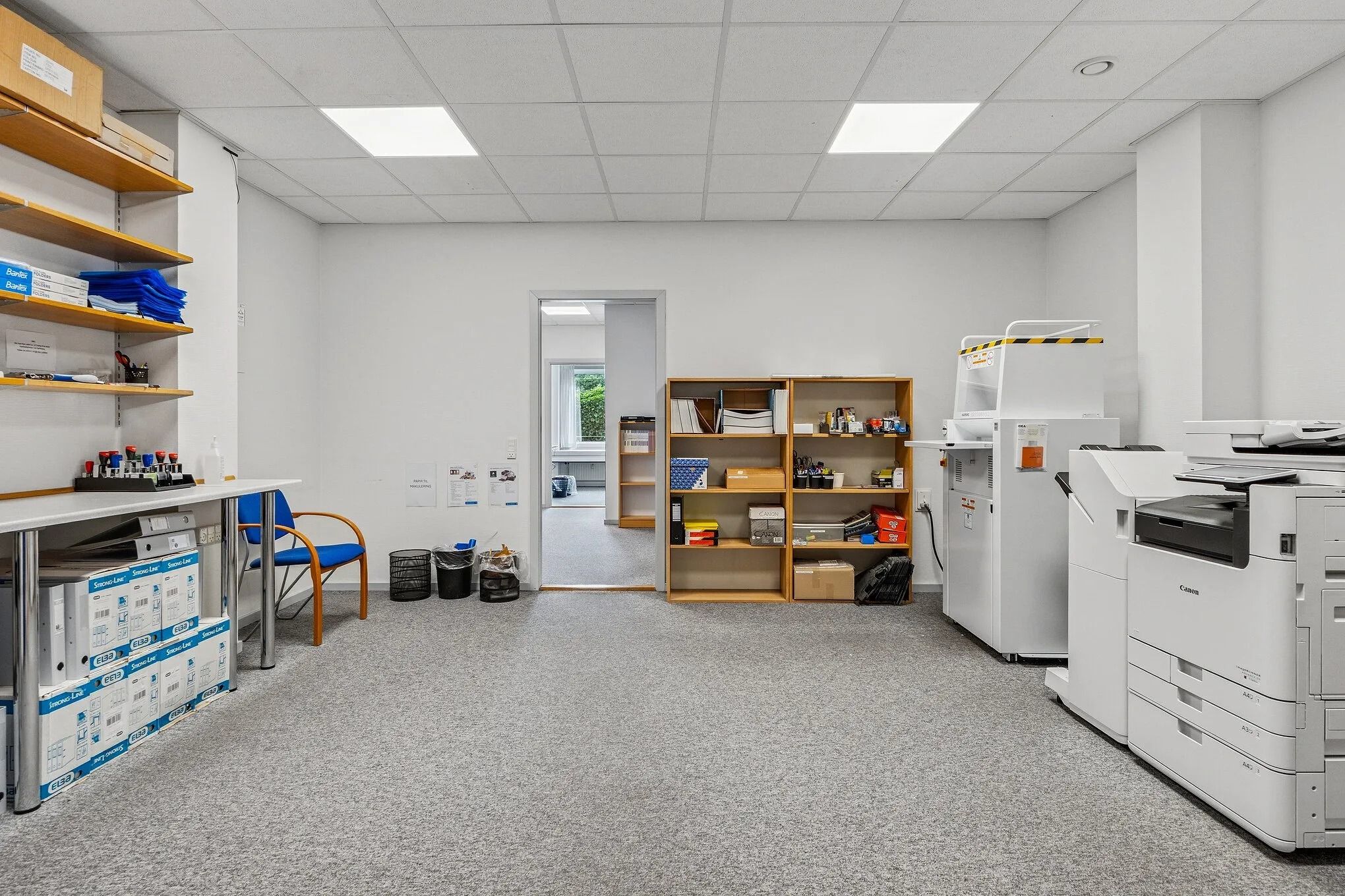 Kontorområde med møbler og teknologi, slanke hvide vægge og lyse loftsbelægninger, tøndekasser og kantoner på hylder og