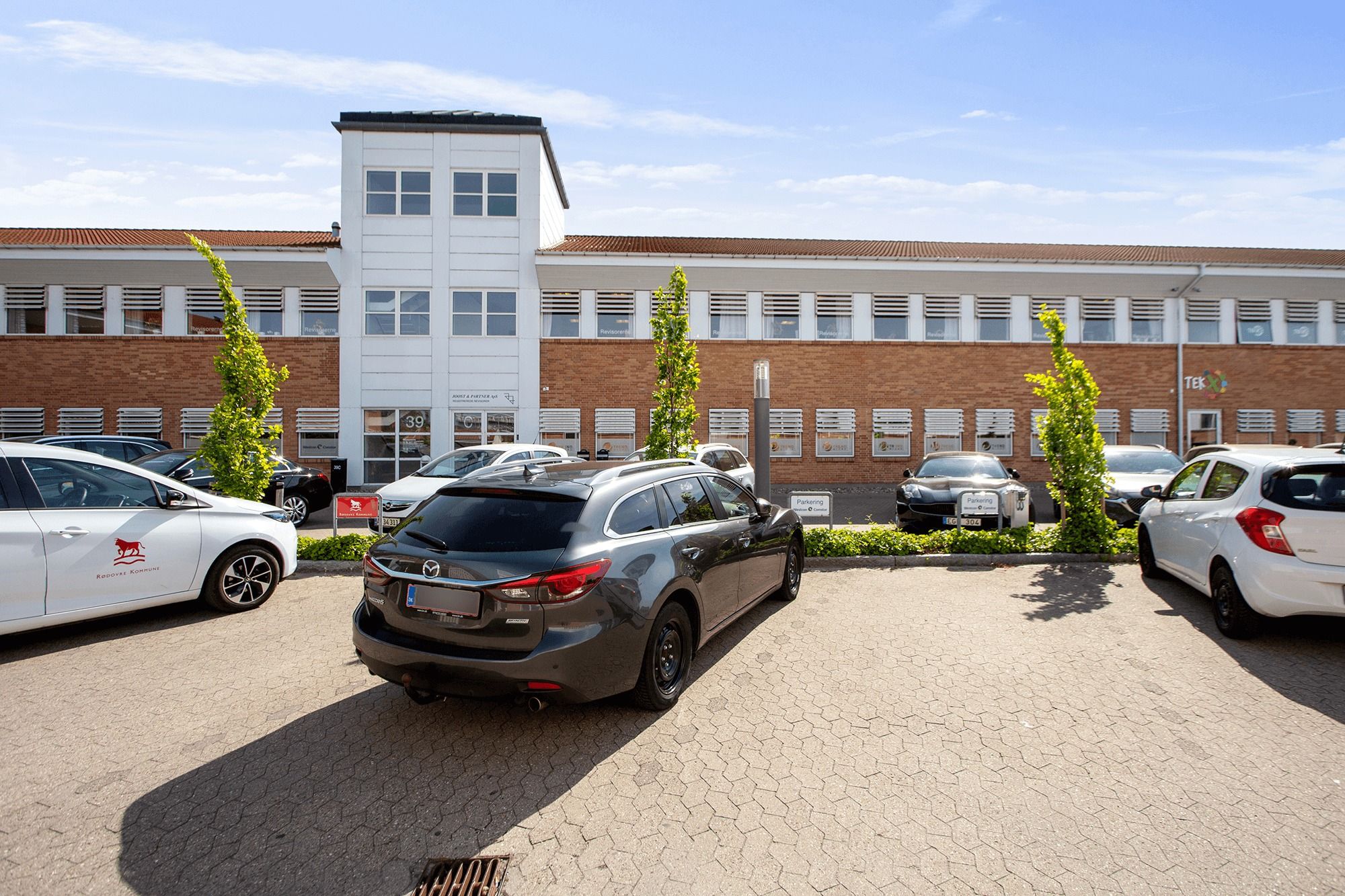 Parkeringsplads med fire parkerede biler og et moderne kontorblok bygget i røde mursten og hvidt materiale