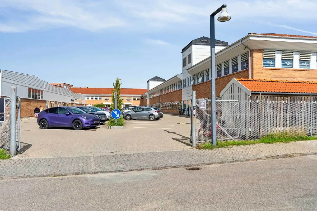 Foran bygningspark med flere faciliteter er parkeringsplads med biler parkeret og en gågade med cykelstativ og gitter.