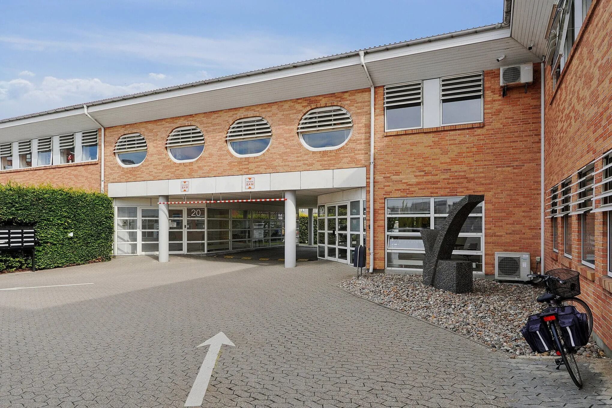 Kontorbyggeri med røde mursten og store runde ruder samt glasindgang. Højre side har en stor skulptur og luftafspiller.