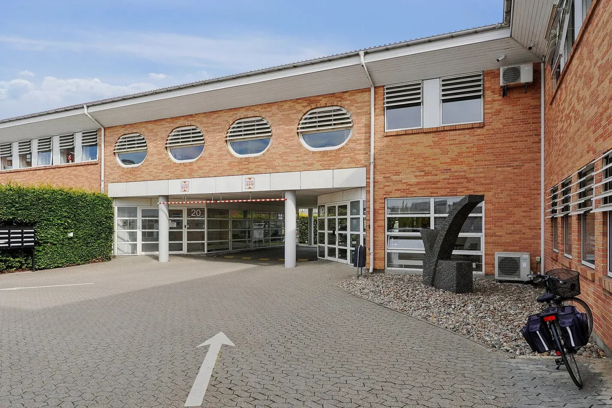 Kontorbyggeri med røde mursten og store runde ruder samt glasindgang. Højre side har en stor skulptur og luftafspiller.
