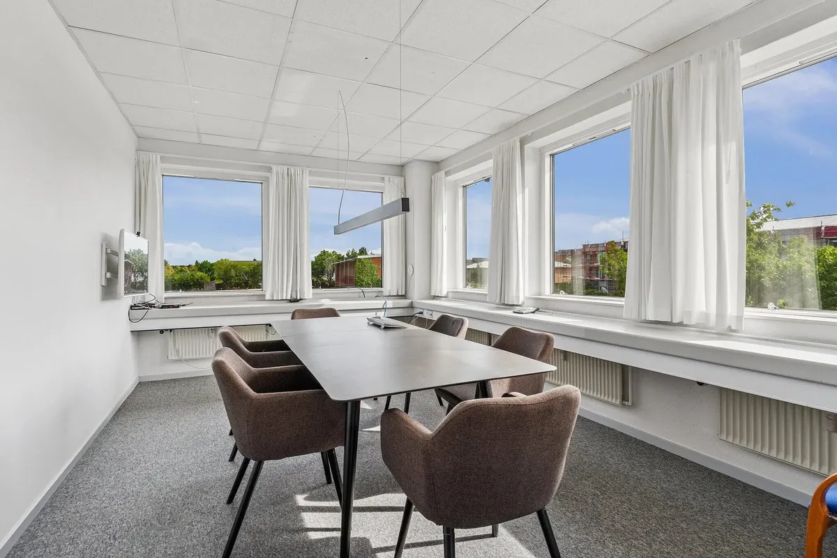 En moderne mødelokale med et langt bord og flere stole, hvor vinduer med hvide gardiner tillader indstrålet af lys og udsigt