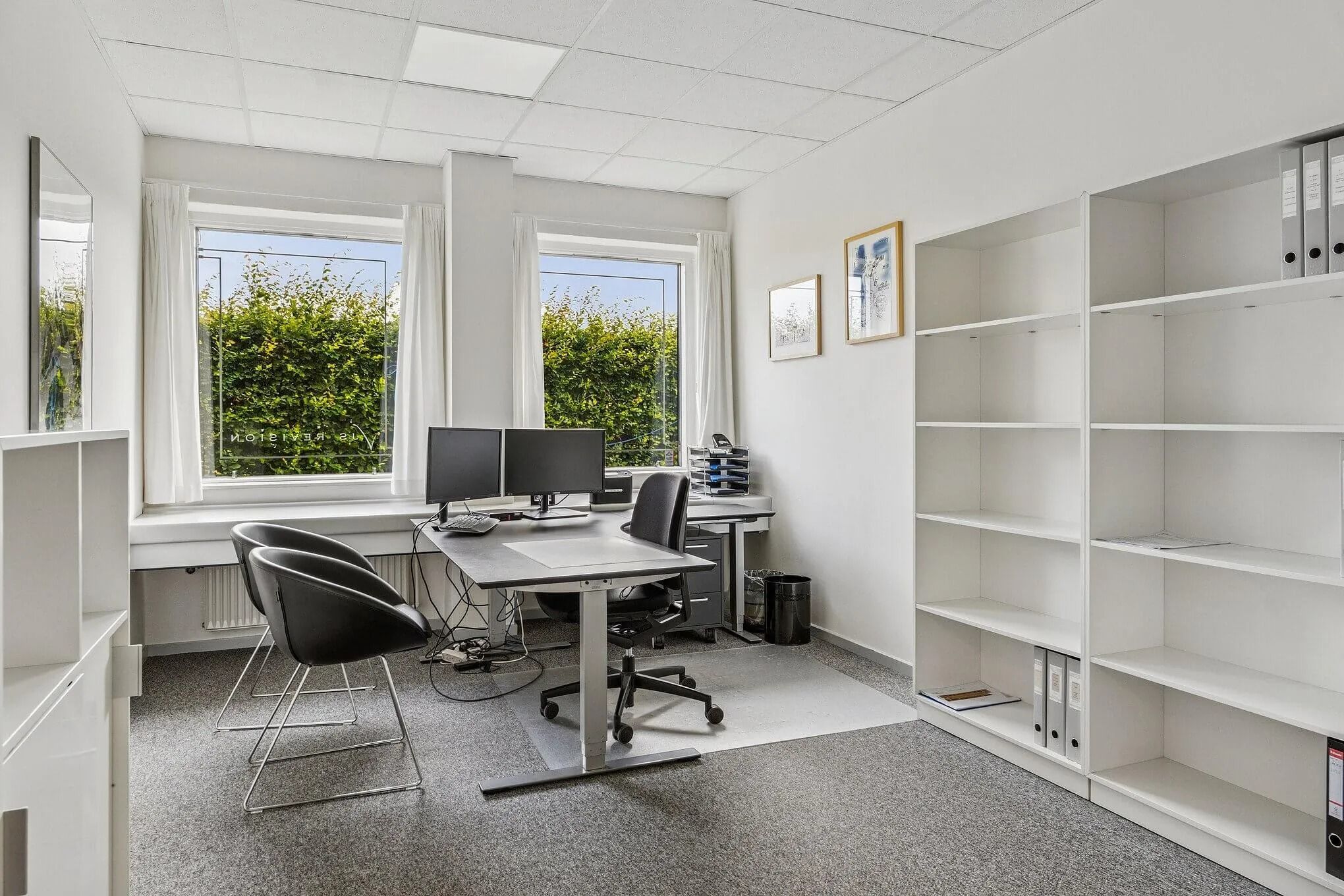 Kontorrum med skrivebord, to skærme, to stole, bibliotek og vindue med udsigt til haven udenfor