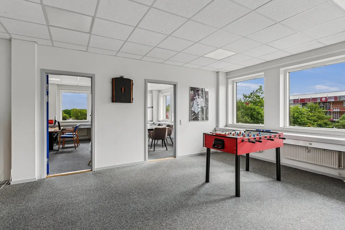 Kontorrum med møbler og et spilbord med bolde, samt vinduer med udsigt til park og bygninger