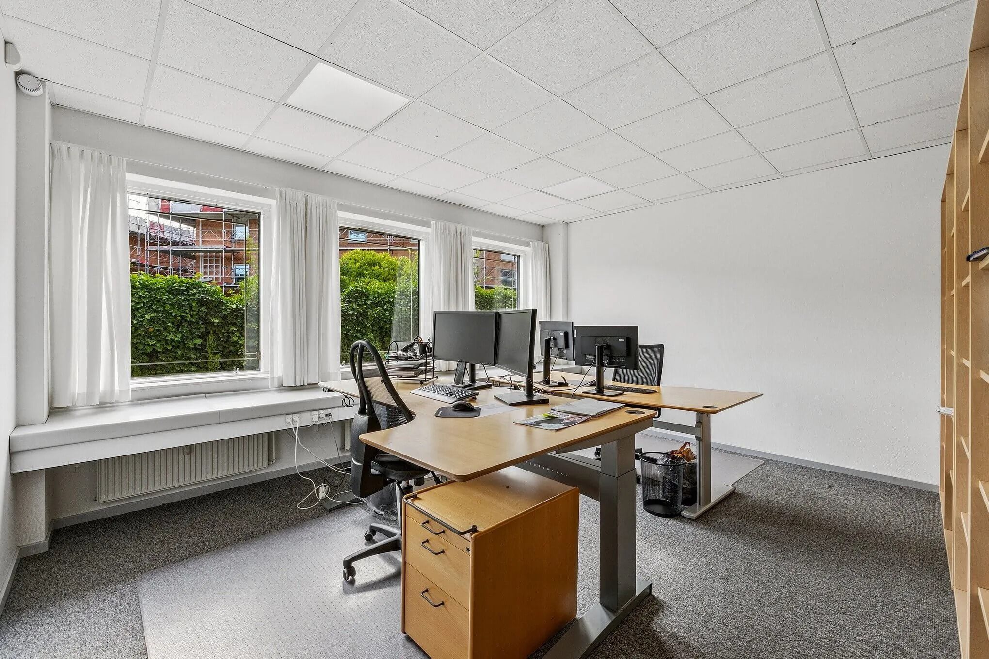 Kontor med skriveborde, stole, computere og vinduer med gardiner, der giver masser af naturlys og udsigt til grønne planter