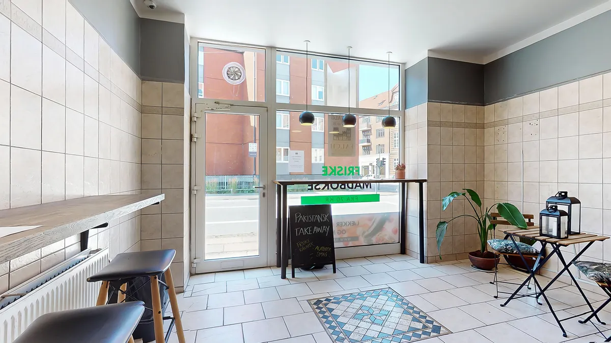 Inde i en lukket restaurant med vinduer og plante. Der er to træborde med lanterner og to stolpe. Der er en vægmaleri og en