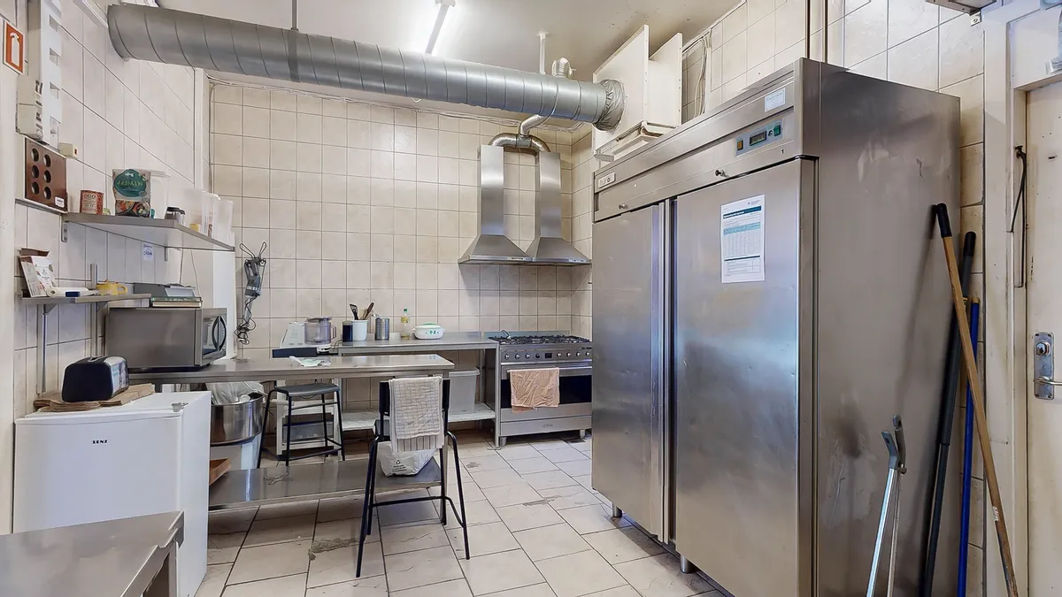 Køkken med flere apparater og hætte over komfuret, samt flere køleskabe og et lille højskab med forskellige genstande