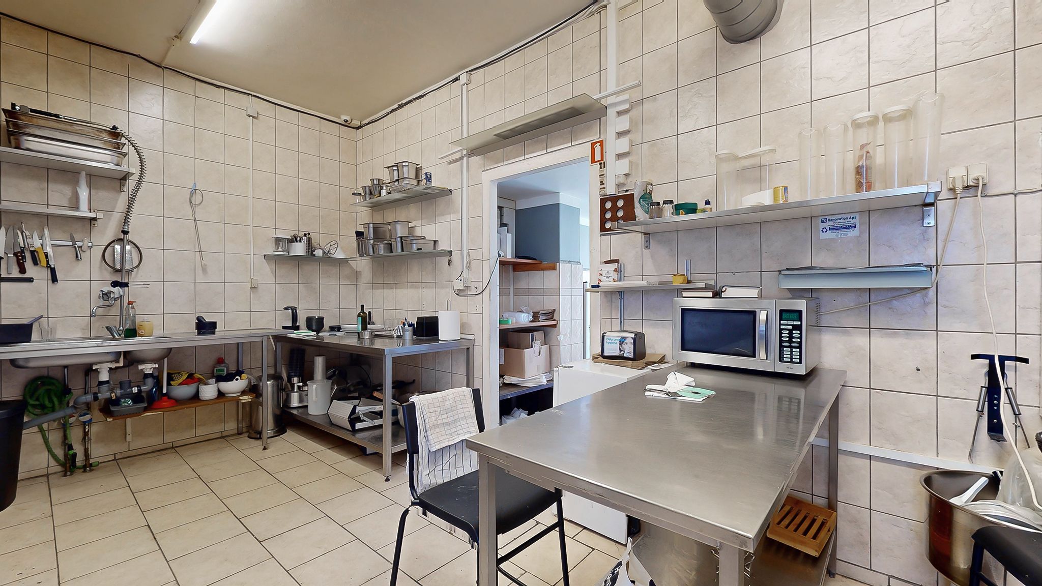 Køkken med metalbord, to stålhylder med diverse køkkenredskaber og en mikroovn og en barbermaskine på den ene hylde