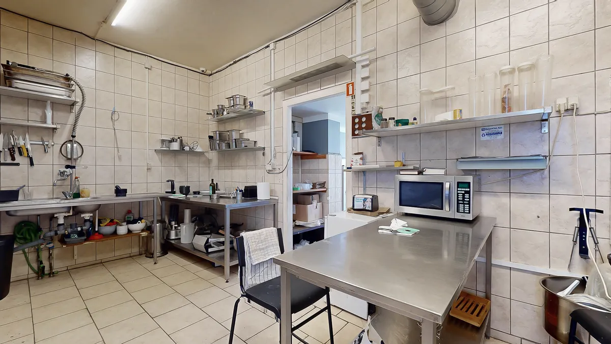 Køkken med metalbord, to stålhylder med diverse køkkenredskaber og en mikroovn og en barbermaskine på den ene hylde