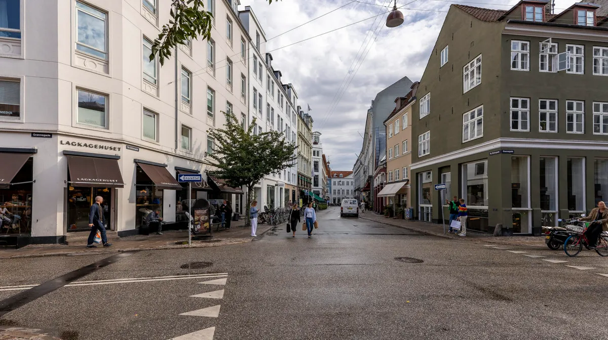 Mennesker går rundt i en gågade med bygninger, træer og vejvisningsskilte